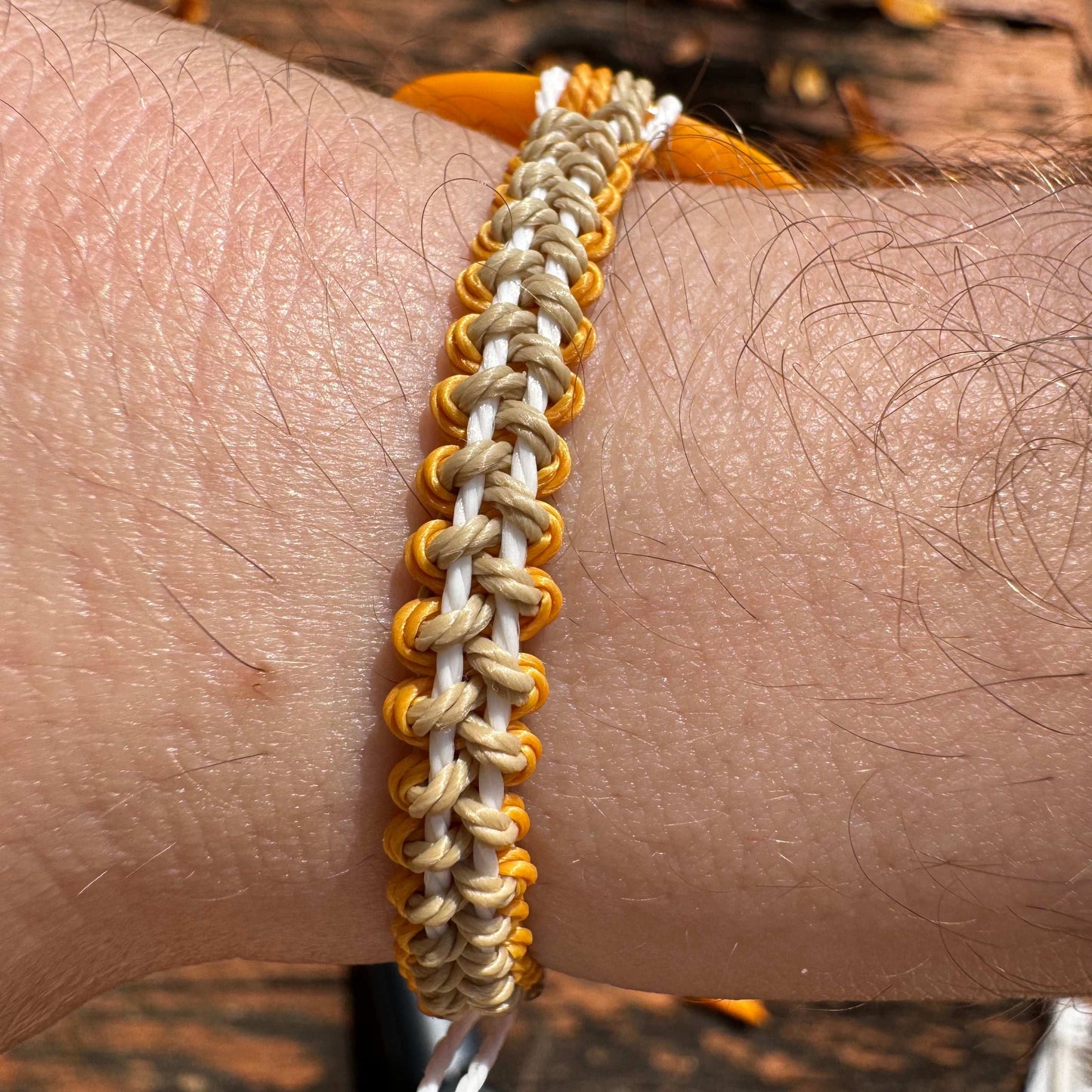 Not Just North picture of the Golden Embrace bracelet. It has a golden yellow circular abalone shell in the center and waxed nylon cords coming from each side that have been braided together in a macrame pattern in golden orange, beige and white threads.