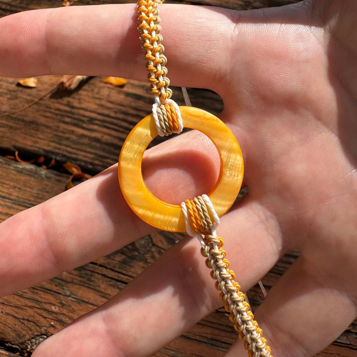 Not Just North picture of the Golden Embrace bracelet. It has a golden yellow circular abalone shell in the center and waxed nylon cords coming from each side that have been braided together in a macrame pattern in golden orange, beige and white threads.