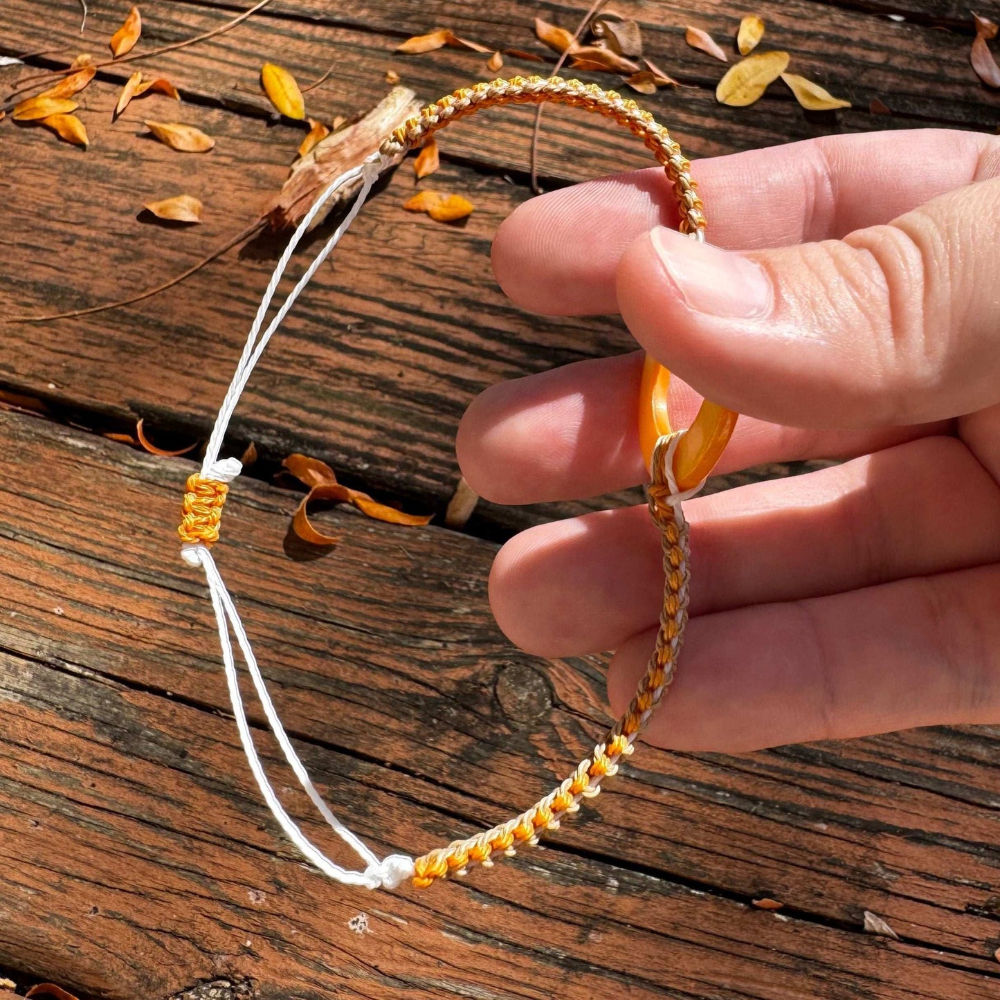 Not Just North picture of the Golden Embrace bracelet. It has a golden yellow circular abalone shell in the center and waxed nylon cords coming from each side that have been braided together in a macrame pattern in golden orange, beige and white threads.