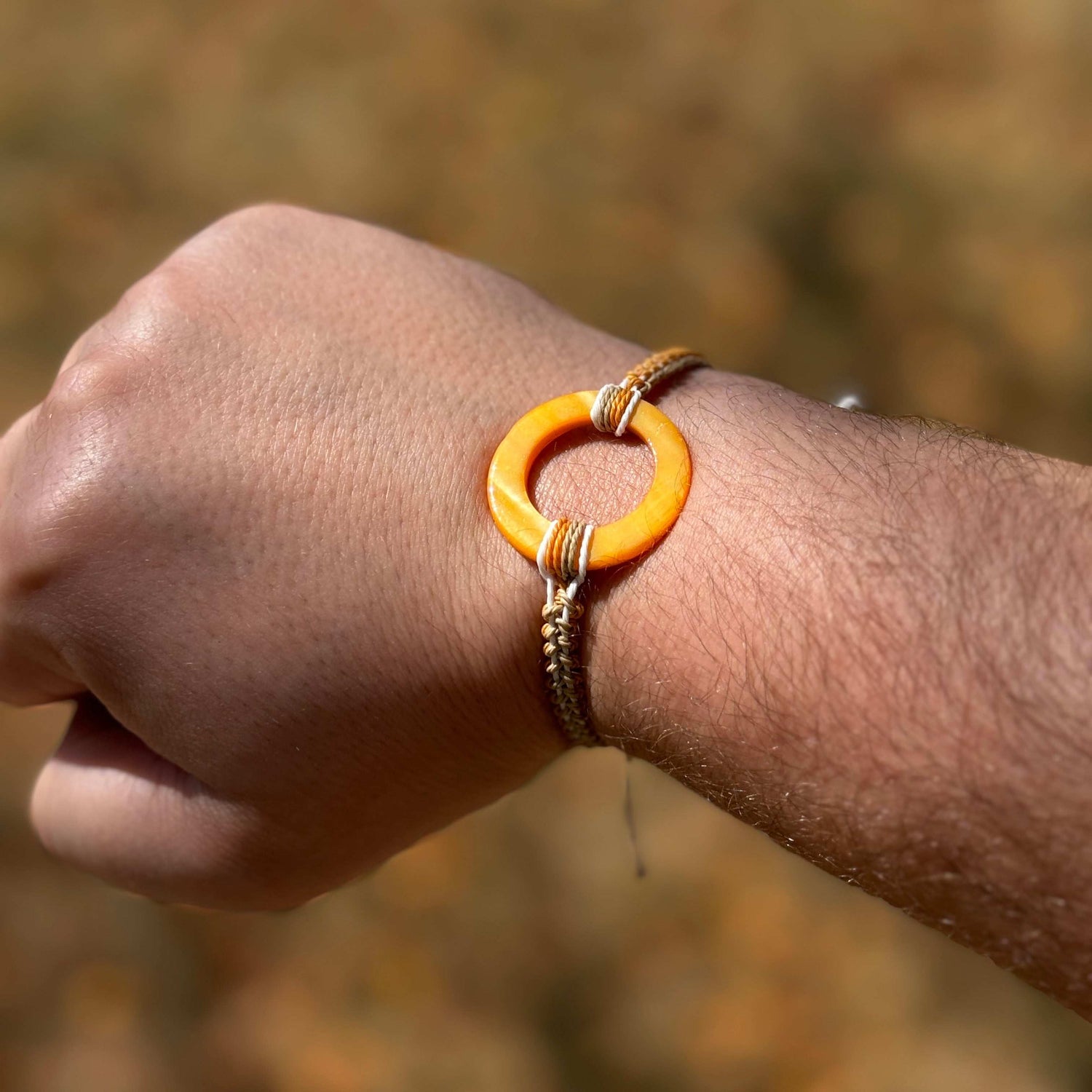 Not Just North picture of the Golden Embrace bracelet. It has a golden yellow circular abalone shell in the center and waxed nylon cords coming from each side that have been braided together in a macrame pattern in golden orange, beige and white threads.
