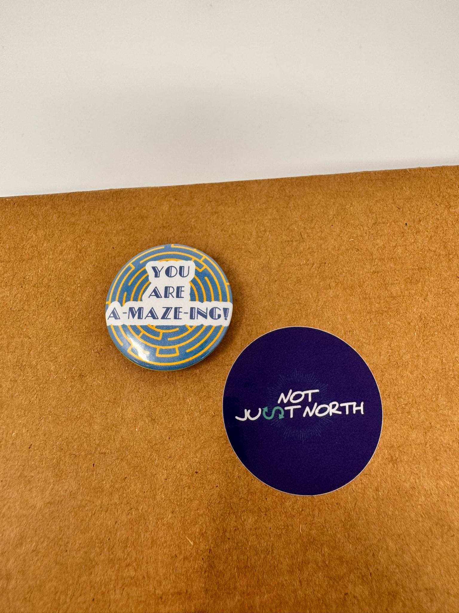 Round button saying amazing sits in the center of the button. A yellow maze on a blue background sit behind the wording. Button is next to a Not Just North Sticker.