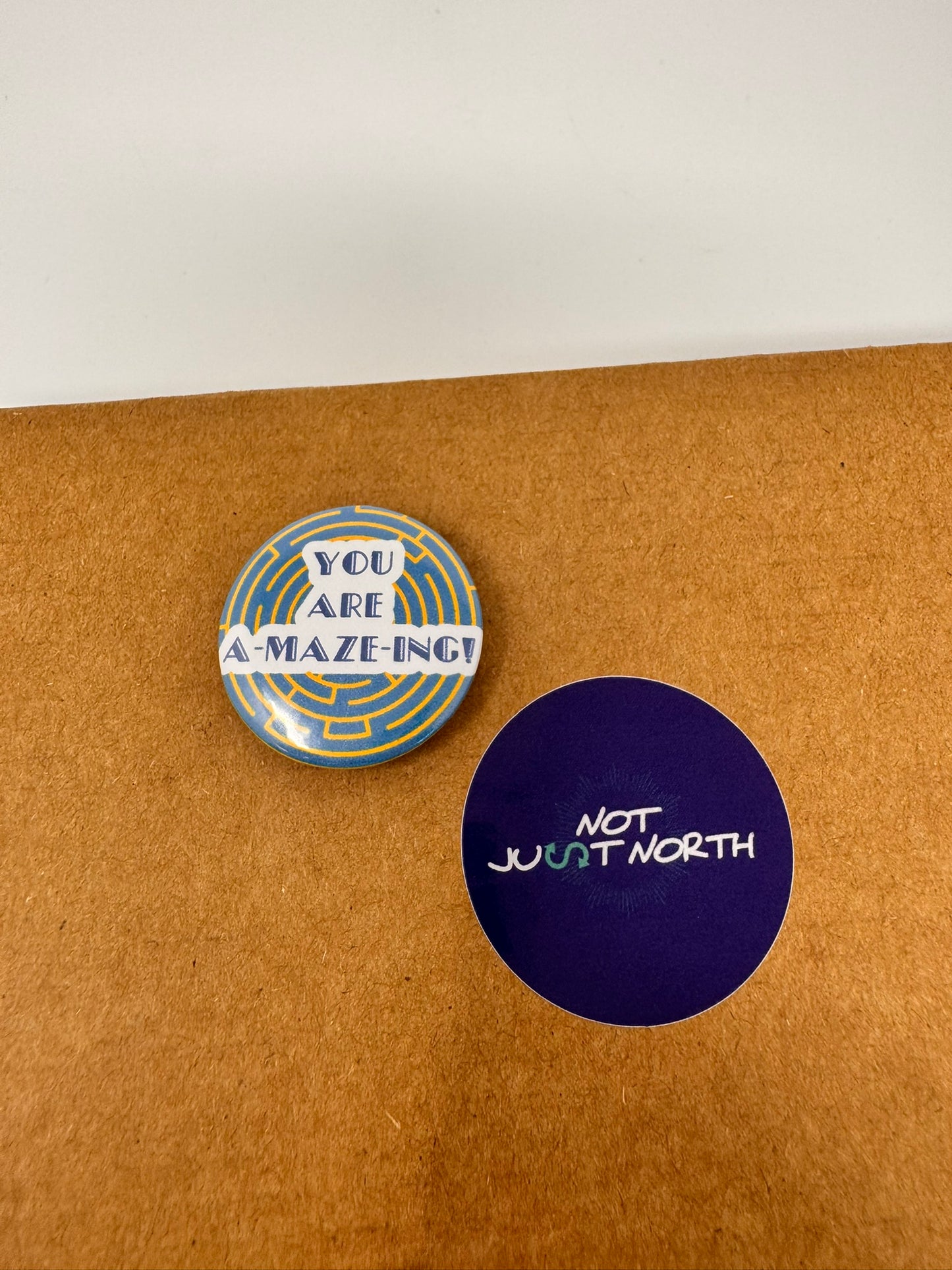 Round button saying amazing sits in the center of the button. A yellow maze on a blue background sit behind the wording. Button is next to a Not Just North Sticker.