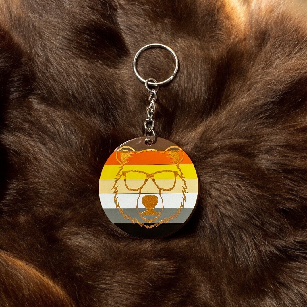 Round acrylic keychain with the gay bear flag in striped colors of black, brown, beige, yellow, cream, and white in the background. Copper bear wearing sunglasses adorns this keychain.