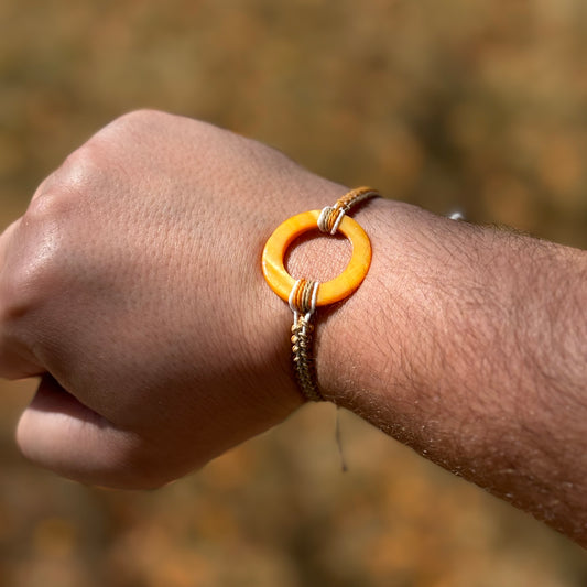 Not Just North picture of the Golden Embrace bracelet. It has a golden yellow circular abalone shell in the center and waxed nylon cords coming from each side that have been braided together in a macrame pattern in golden orange, beige and white threads.