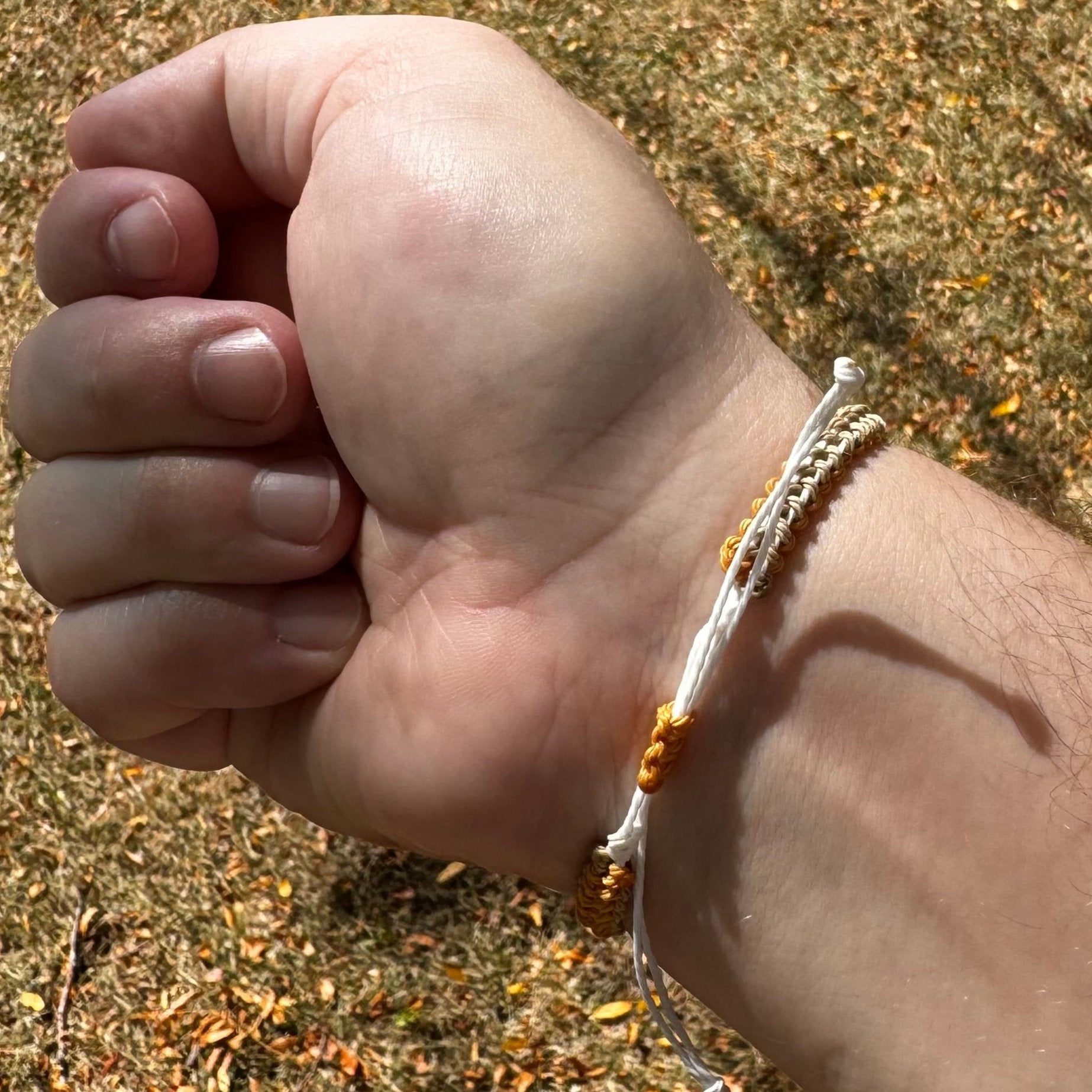 Not Just North picture of the Golden Embrace bracelet. It has a golden yellow circular abalone shell in the center and waxed nylon cords coming from each side that have been braided together in a macrame pattern in golden orange, beige and white threads.