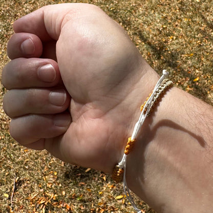 Not Just North picture of the Golden Embrace bracelet. It has a golden yellow circular abalone shell in the center and waxed nylon cords coming from each side that have been braided together in a macrame pattern in golden orange, beige and white threads.