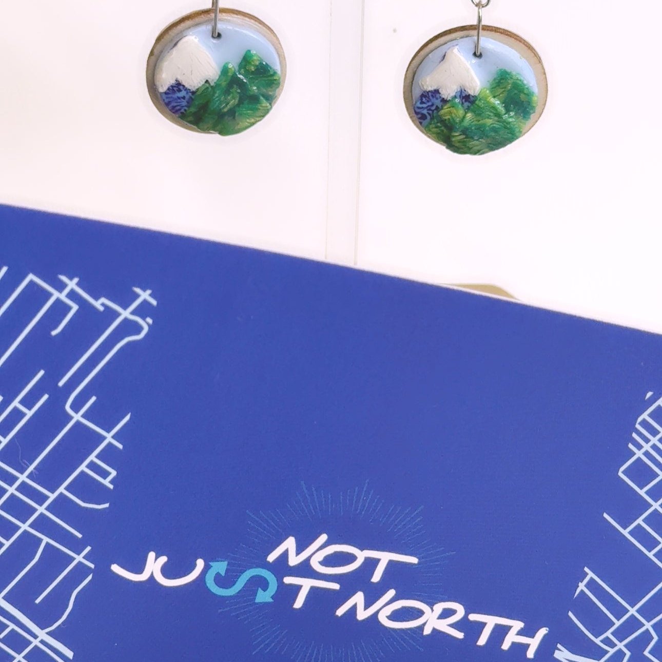 Not Just North Rise Up Earrings White Snowcapped Purple Mountains with Green Pine Trees in the Foreground. Contains the brand name Not Just North