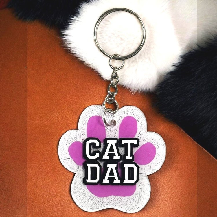paw shaped keychain with white background that has been scratched to give the effect of fur. White letters that say cat dad with black border.
