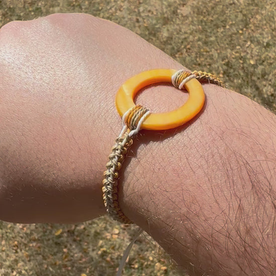 Not Just North video of the Golden Embrace bracelet. It has a golden yellow circular abalone shell in the center and waxed nylon cords coming from each side that have been braided together in a macrame pattern in golden orange, beige and white threads.