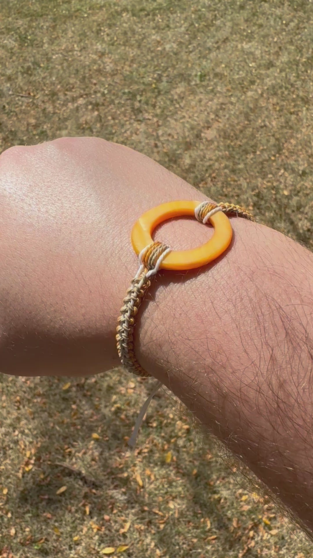 Not Just North video of the Golden Embrace bracelet. It has a golden yellow circular abalone shell in the center and waxed nylon cords coming from each side that have been braided together in a macrame pattern in golden orange, beige and white threads.